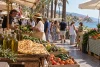 Un marché méditerranéen en bord de mer avec des étals de légumes, de focaccia et d’huile d’olive.